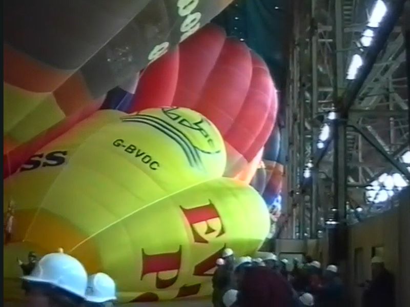 A colour picture of several hot air balloons being inflated inside an airship hangar